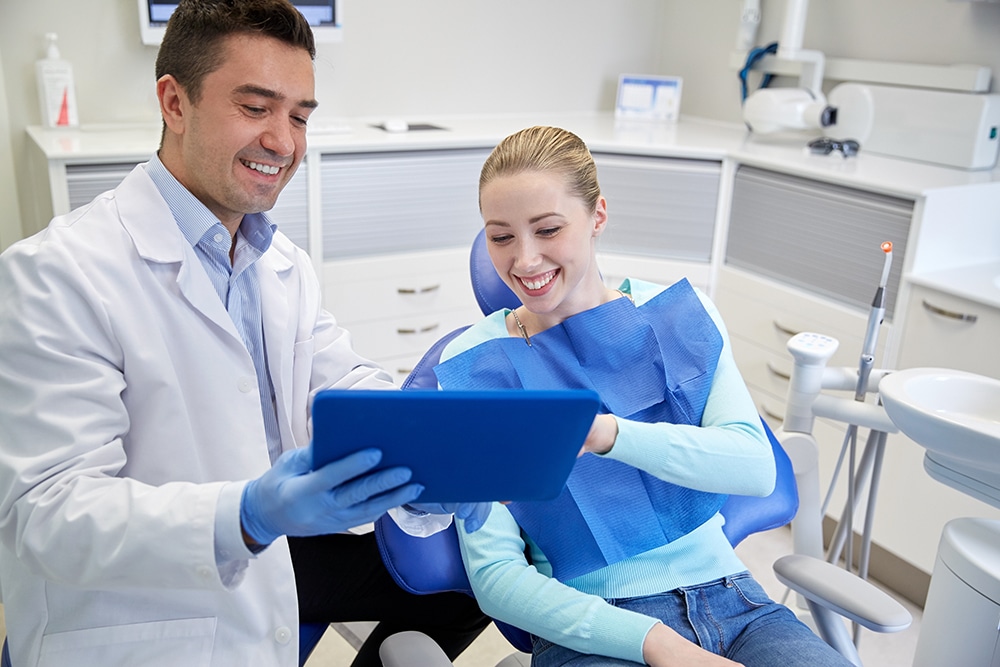 Dentist showing treatment information on a tablet to a patient seated in a dental chair with a laser dental device visible nearby.
